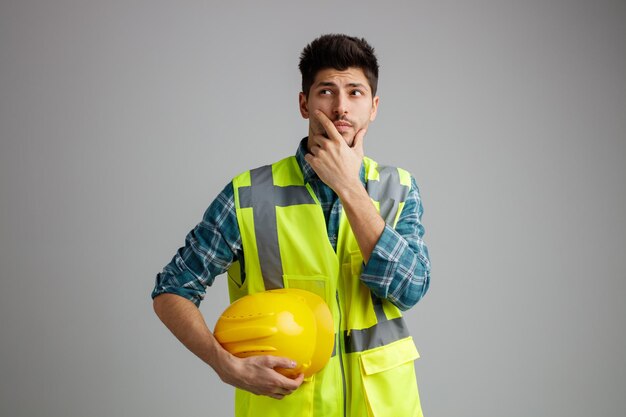 Onzekere jonge mannelijke ingenieur die een uniform draagt en naar de zijkant kijkt terwijl hij een veiligheidshelm vasthoudt terwijl hij de hand op de kin houdt geïsoleerd op een witte achtergrond