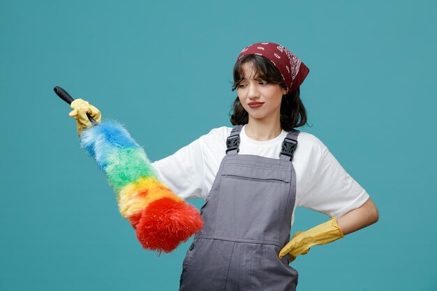 onzekere jonge vrouwelijke schoonmaker die uniforme bandana en rubberen handschoenen draagt en naar plumeau kijkt terwijl ze de hand op de taille houdt geïsoleerd op blauwe achtergrond