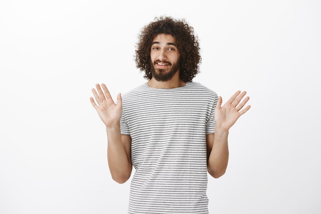 Onzorgvuldig knap mannelijk model met afrokapsel en zieke baard, palmen in overgave opheffen en nerveus glimlachen