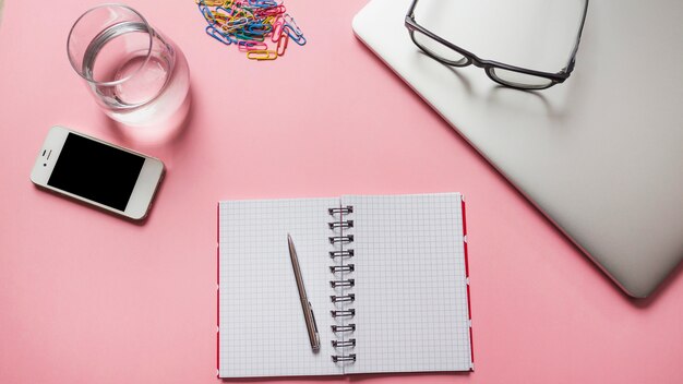 Oogglazen op laptop met kantoorbehoeften; smartphone en een glas water op roze achtergrond