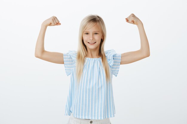 Op een dag wordt een meisje een beroemde sportvrouw. Kleine zelfverzekerde jongen met blond haar in blauwe blouse, handen opheffen met gebalde vuisten, spieren tonen, glimlachend van tevredenheid, gelukkig zijn met eigen kracht