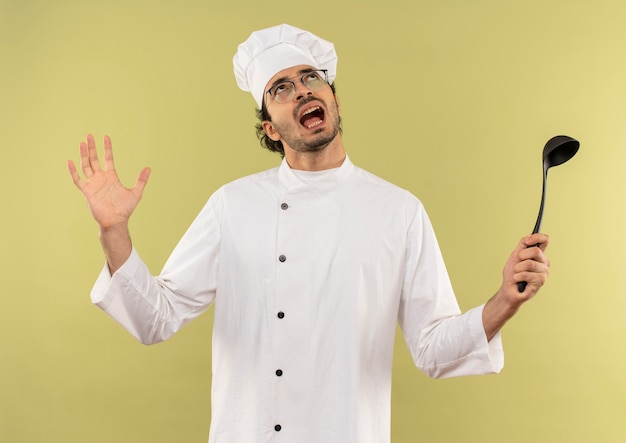 Op zoek naar boze jonge mannelijke kok dragen uniform van de chef-kok en glazen pollepel houden en spreidde hand