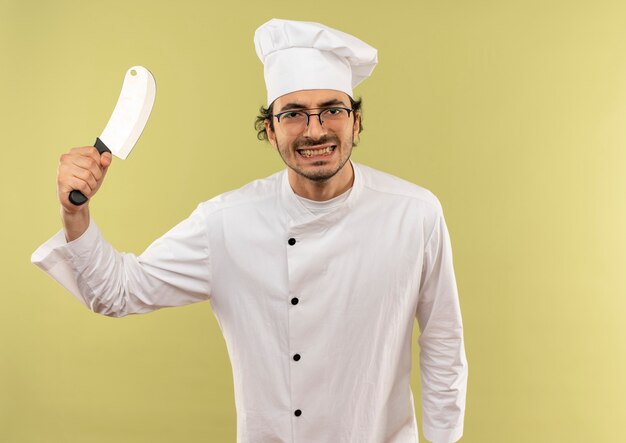 Op zoek naar een tevreden jonge mannelijke kok met een uniform van de chef-kok en een bril die een hakmes op een groene achtergrond opheft?