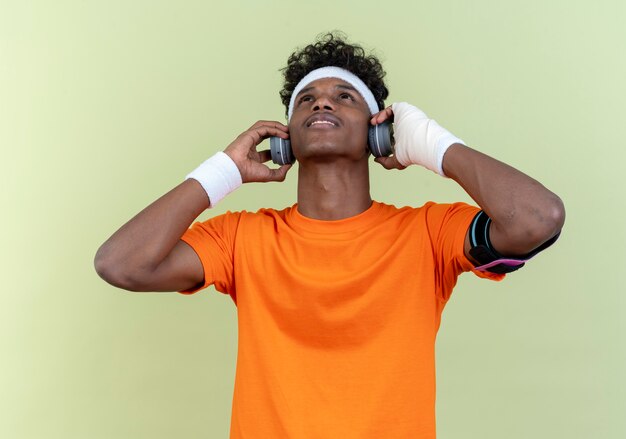 Op zoek naar jonge afro-amerikaanse sportieve man met hoofdband en polsbandje en telefoonarmband met koptelefoon geïsoleerd op groene achtergrond