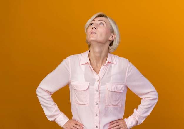 Op zoek naar jonge blonde Slavische vrouw hand op heup zetten geïsoleerd op een oranje achtergrond