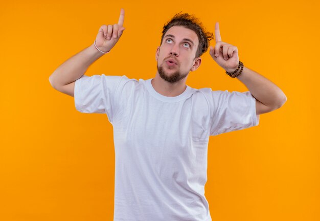 Op zoek naar jonge man met een wit t-shirt wijst naar omhoog op geïsoleerde oranje achtergrond