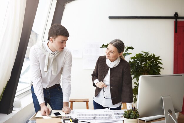 Openhartig shot van twee Europese architecten die een discussie voeren op kantoor, aan het bureau staan met computer, tekeningen en gereedschappen, glimlachend naar elkaar, tevreden met gemeenschappelijk werk. Mensen en samenwerking