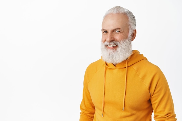 Openhartige gelukkige oude man met lange stijlvolle baard, lachen en glimlachen, er gelukkig uitzien, moderne oranje hoodie dragen, staande over witte muur