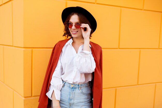 Openlucht dicht omhooggaand portret van het zalige donkerbruine kortharige vrouwelijke stellen over gele muur. Trendy hoed, roze bril, witte blouse en jeans.