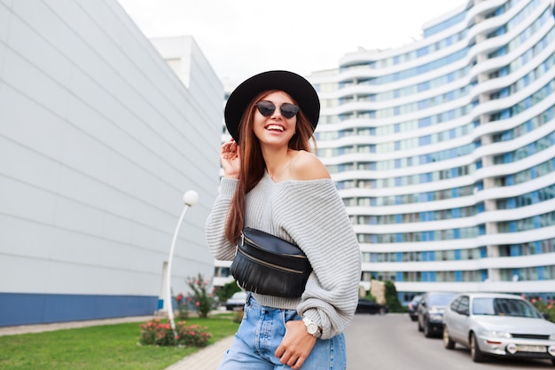 Openluchtbeeld van blij meisje in zwarte wollen hoed en grijze dalingssweater die en van gang in moderne stedelijke stad springen genieten.