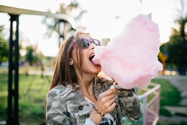 Openluchtfoto van een jonge trendy vrouw die in zonnebril een gesponnen suiker eet