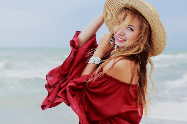 Openluchtlevensstijlportret van vrij witte gembervrouw in het modieuze kleding stellen op het strand dichtbij oceaan. blu hemel. winderig weer.