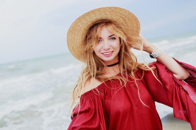 Openluchtlevensstijlportret van vrij witte gembervrouw in het modieuze kleding stellen op het strand dichtbij oceaan. Blu hemel. Winderig weer.