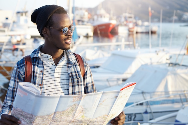 Openluchtportret van de afrikaanse mens die gelukkig vóór reis kijkt, wachtend op zijn vrienden in haven, houdend kaartkaart, opgewekt en blij voelt, anticiperend op avonturen, plaatsen en goede ervaring