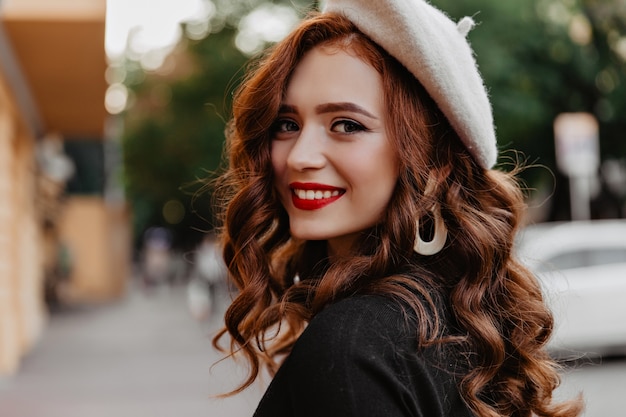 Openluchtportret van lachende gembervrouw met rode lippen die van de herfstochtend genieten. debonair franse dame in baret terugkijkend.