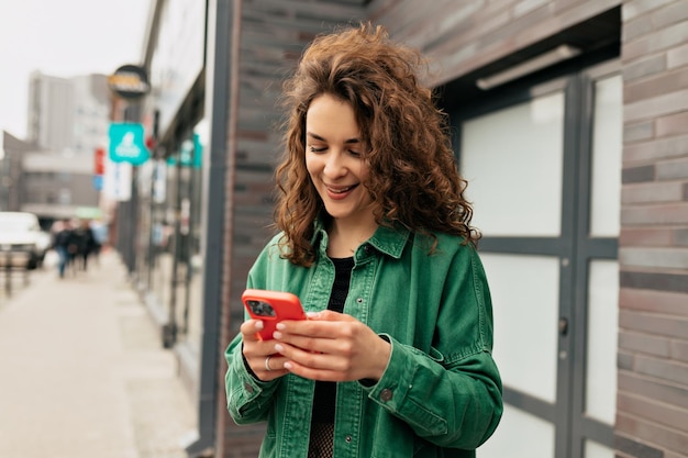 Openluchtportret van mooi stijlvol meisje met krullen die groen overhemd dragen die smartphone met glimlach gebruiken Zorgeloos jong Kaukasisch meisje gebruikt moderne smartphone die zich buitenshuis bevindt