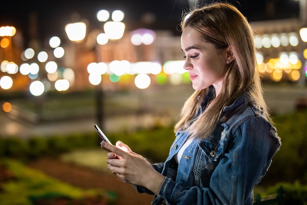 Openluchtportret van mooie jonge vrouw die haar mobiele telefoon bij nacht met behulp van.