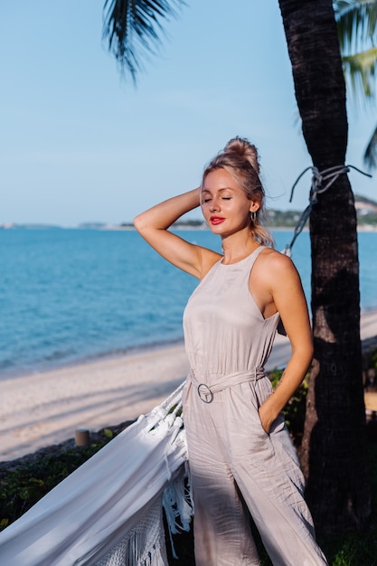 Openluchtportret van rijke Kaukasische vrouw in klassieke jumpsuit met rode lippenstift door hangmat op vakantie buiten villahotel