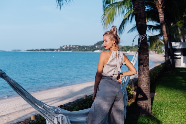 Openluchtportret van rijke Kaukasische vrouw in klassieke jumpsuit met rode lippenstift door hangmat op vakantie buiten villahotel