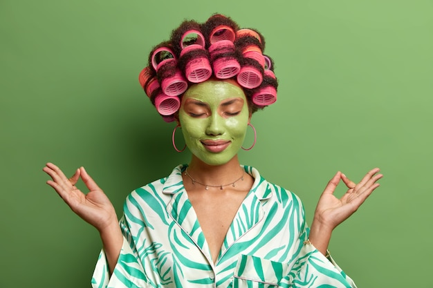 Opgelucht etnische vrouw draagt een gezichtsmasker van de schoonheid, staat met gesloten ogen, mediteert thuis terwijl ze spabehandelingen ondergaat, yoga traint, perfecte haarstijl maakt, ontspant in huiselijke sfeer.