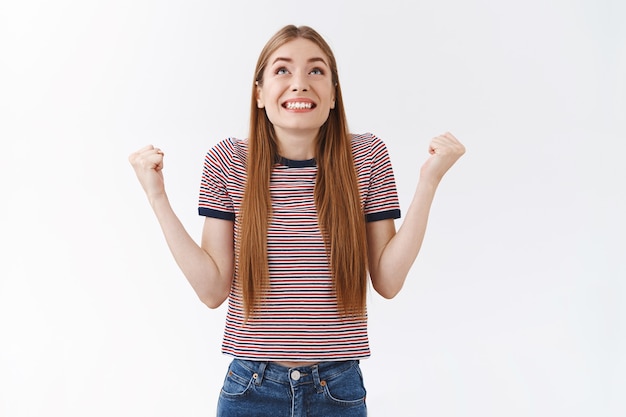 Opgeluchte, gelukkige knappe vrouw in gestreept t-shirt godzijdank en vuist pompt vrolijk, kijk vrolijk glimlachend naar de lucht, triomfeert van geweldig nieuws, staat blij en viert de overwinning