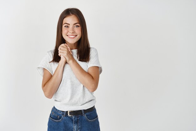 Opgetogen jonge vrouw die met dankbaarheid en geluk kijkt, de handen in elkaar grijpt en over een witte muur staat