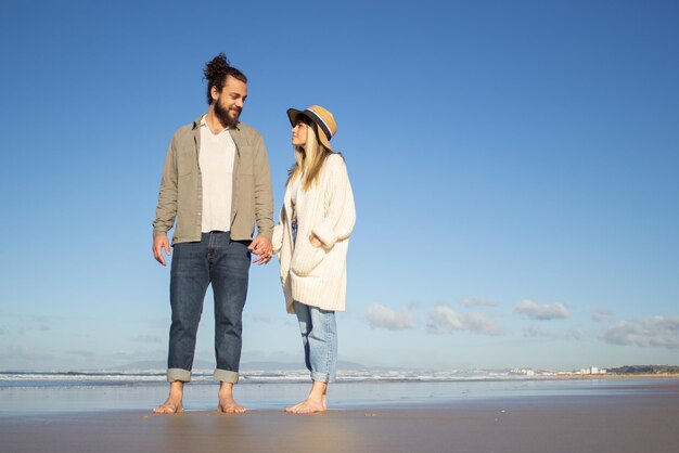 Opgetogen paar die op strand lopen. Bebaarde man en vrouw in vrijetijdskleding kijken elkaar aan, hand in hand. Liefde, vakantie, genegenheid concept