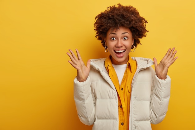 Gratis foto opgewekt verrast vrouw met donkere huid heft handpalmen op, blij om een mooi cadeau te krijgen, draagt witte warme winterjas, gele jas, drukt goede emoties uit, glimlacht breed, heeft een blije reactie op verrassing