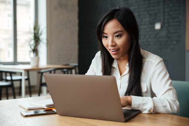 Opgewekte jonge Aziatische onderneemster die laptop computer met behulp van