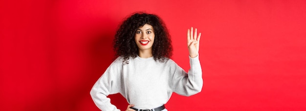 Gratis foto opgewekte vrouw met krullend haar die nummer vier toont met vingers die orde maken die zich tegen rode bac bevinden