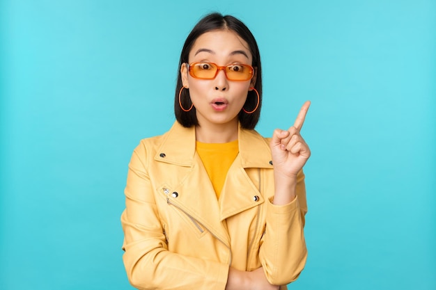 Opgewonden Aziatisch meisje dat vinger opsteekt en suggereert dat een eureka-teken een plan heeft dat in een zonnebril op een blauwe achtergrond staat