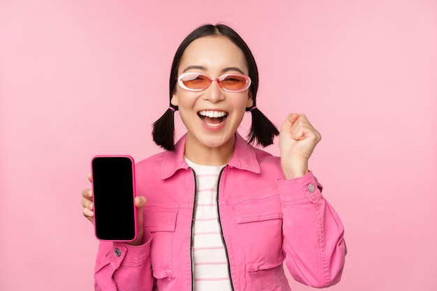Opgewonden Aziatisch meisje lacht en glimlacht en toont een smartphone-applicatie op het scherm van een mobiele telefoon die over een roze achtergrond staat