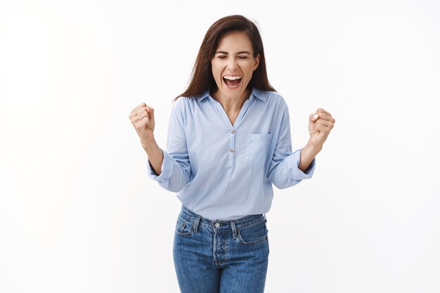 Opgewonden bemoedigde, gemotiveerde knappe volwassen vrouw schreeuw hoera ja, vuist pomp vreugdevol, vier overwinning, triomfeert vrolijk, sluit ogen die vrolijk schreeuwen, sta op witte muur