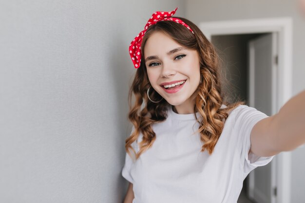 Opgewonden blanke vrouw in oorbellen selfie maken in de ochtend. Indoor foto van zalige stijlvolle dame draagt lint in haar.