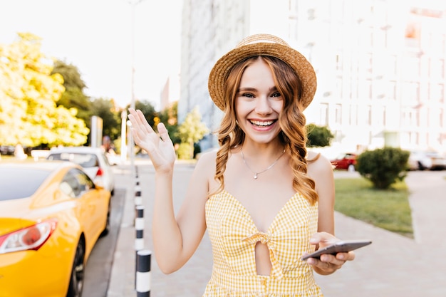 Opgewonden blanke vrouw zwaaiende hand tijdens het poseren op straat in de ochtend. Blond meisje met krullend kapsel gele jurk dragen in zomerdag.