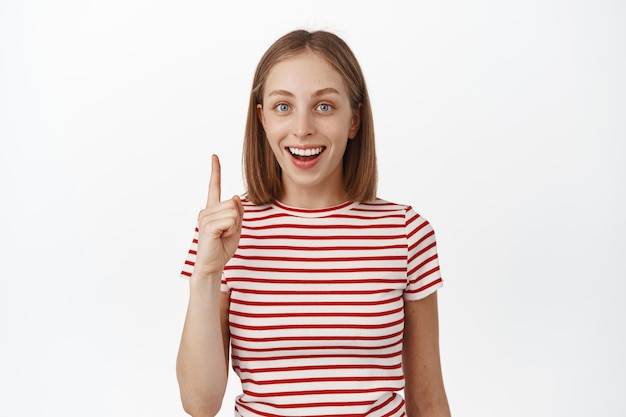 Opgewonden blond meisje dat witte tanden glimlacht, vinger omhoog wijst, een idee kreeg, iets interessants suggereert, banner of advertentie hieronder toont, staande in gestreept t-shirt op witte achtergrond.