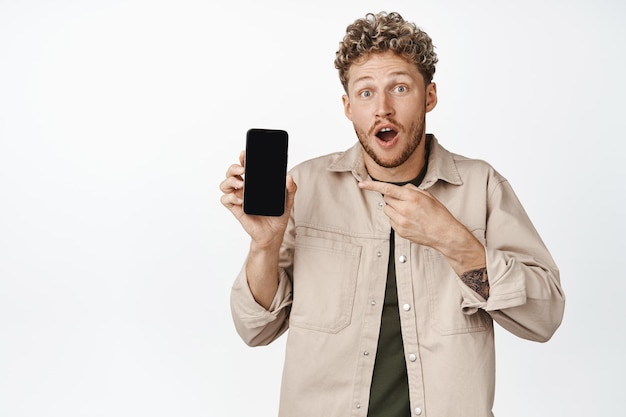 Opgewonden blonde krullende man die met de vinger naar het scherm van de mobiele telefoon wijst en er verbaasd uitziet met geweldig nieuws online op een witte achtergrond