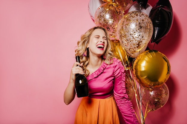 Opgewonden blonde vrouw met champagne die op roze muur lacht