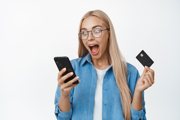 Opgewonden blonde vrouw met een bril die naar het scherm van de smartphone kijkt en schreeuwt, verrast en verbaasd op wit staart