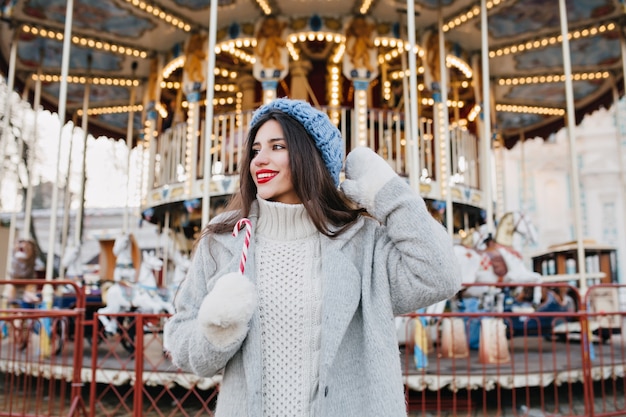 Opgewonden brunette meisje in onscherpte gebreide muts te wachten op vriend in pretpark in winterdag. Buiten foto van gelukkige vrouw met donker haar riet van het suikergoed houden en poseren voor carrousel.