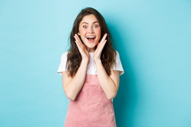 Opgewonden brunette vrouw met schattig krullend kapsel, hijgend verbaasd en verbaasd, kijk verbaasd naar de camera, win of ontvang een verrassingsgeschenk, staande over blauwe achtergrond.