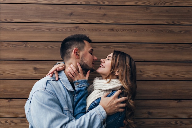 Opgewonden donkerharige man die zijn geliefde vrouw omhelst. Elegante mooi meisje kussen vriendje op houten muur.