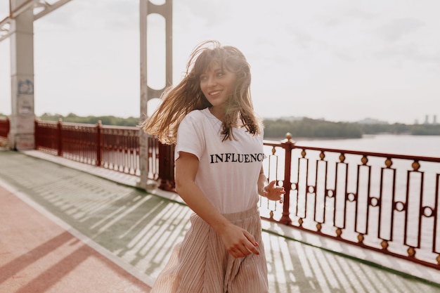 Opgewonden emotioneel meisje met los vliegend haar in een wit t-shirt en een losse roze rok die in de ochtendzon op de brug springt