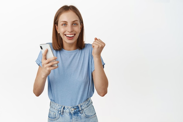Opgewonden en gelukkige jonge vrouw die smartphone vasthoudt, geld wint op mobiele telefoon, dagelijks doel in app bereikt, van geluk springt en tevreden glimlacht, witte achtergrond