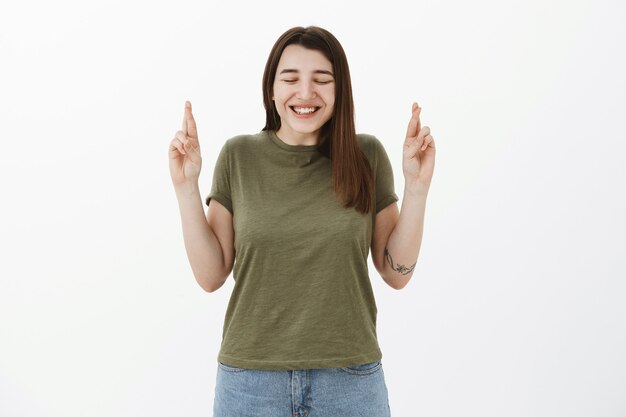 Opgewonden en schattig oprecht jong meisje met tattto die zich bukt van spanning en geluk met gesloten ogen en een blije glimlach, vingers kruisen voor geluk, wensen doen, bidden om droom die uitkomt over grijze muur