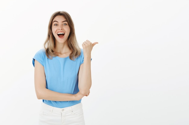 Opgewonden en verbaasd blonde vrouw wijzende duim naar rechts, aankondiging tonen