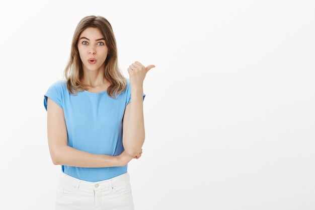 Opgewonden en verbaasd blonde vrouw wijzende duim naar rechts, aankondiging tonen