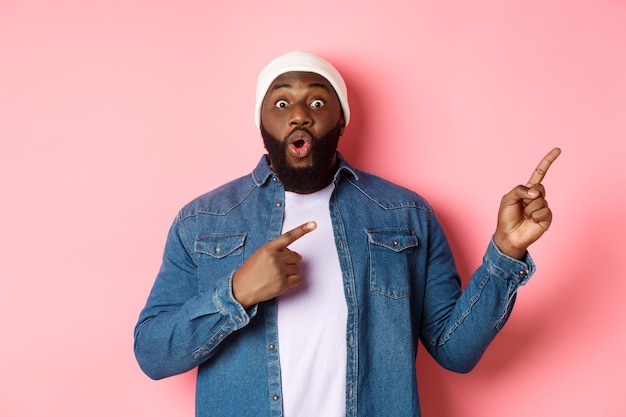 Opgewonden en verbaasde zwarte man die een geweldig aanbod toont, met de vingers naar de kopieerruimte wijst, in een hipsterbeanie en een spijkerblouse op een roze achtergrond staat
