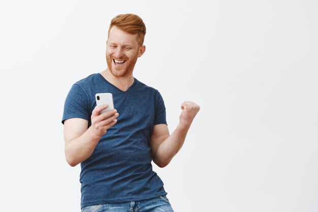 Opgewonden gelukkig en vieren knappe roodharige man met haren, vuist opheffen in overwinning gebaar, smartphone vasthouden