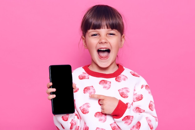 Opgewonden gelukkig lachend vrouwelijk soort met geopende mond mobiele telefoon in handen houden en wijzend op blanco display met wijsvinger, poseren geïsoleerd over roze muur.